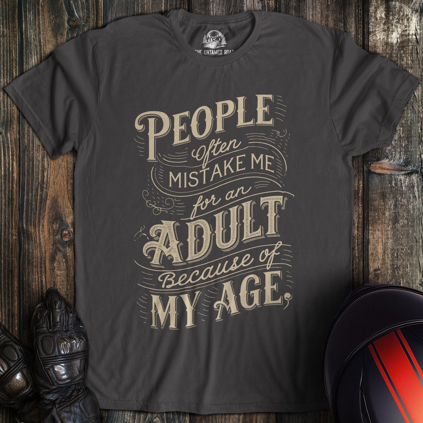 A charcoal colored t-shirt with the words "mistake me for adult" printed on the front.
