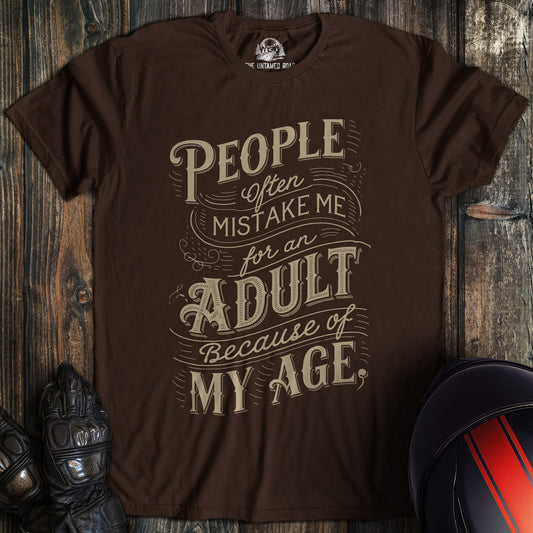 A dark chocolate colored t-shirt with the words "mistake me for adult" printed on the front.