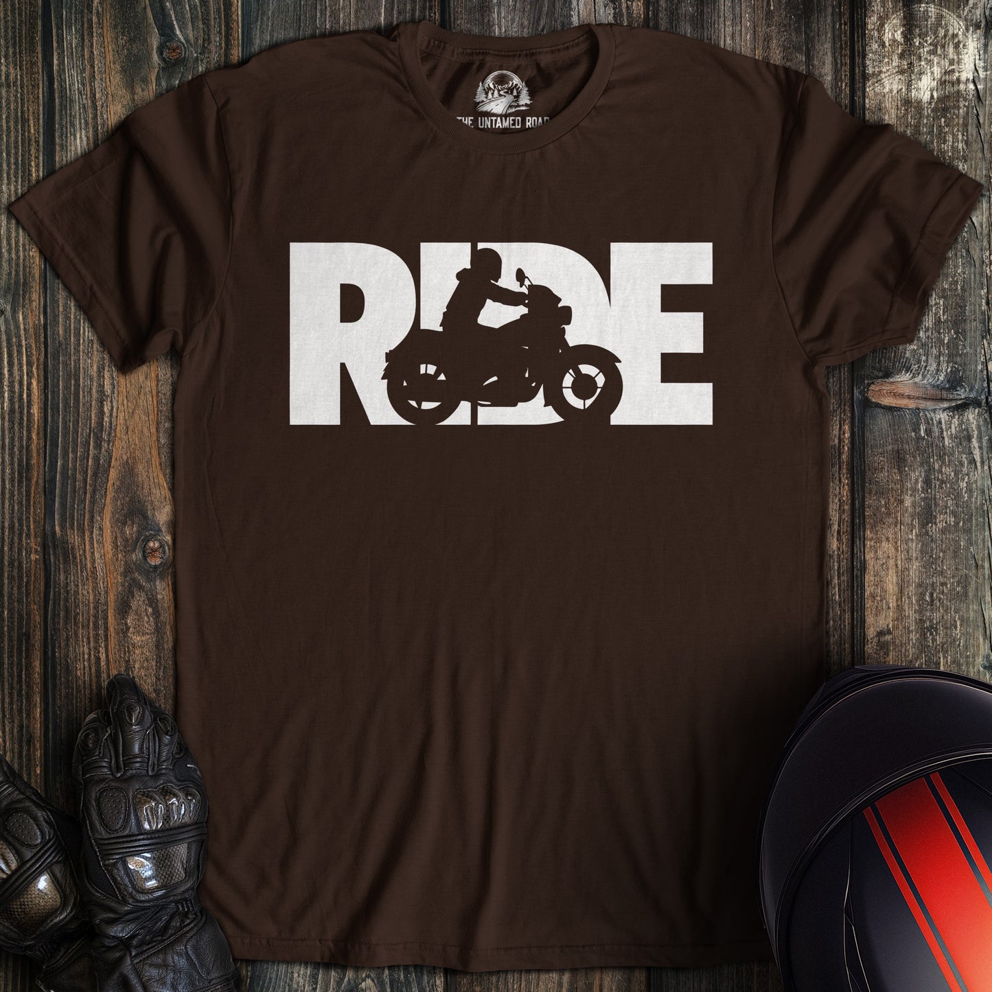 Brown t-shirt with 'RIDE' graphic on a wooden surface with motorcycle gear.