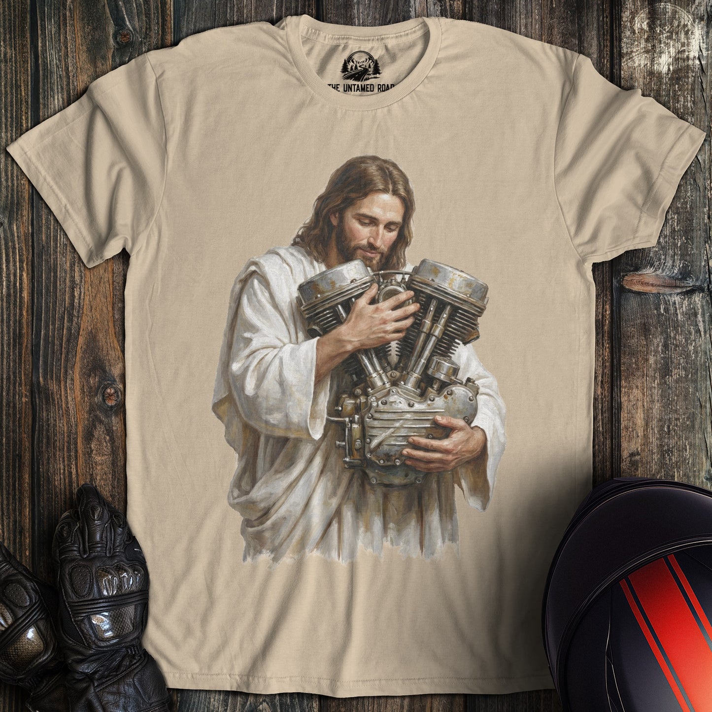 A sand colored t-shirt featuring a funny graphic of Jesus cradling a large motorcycle engine.