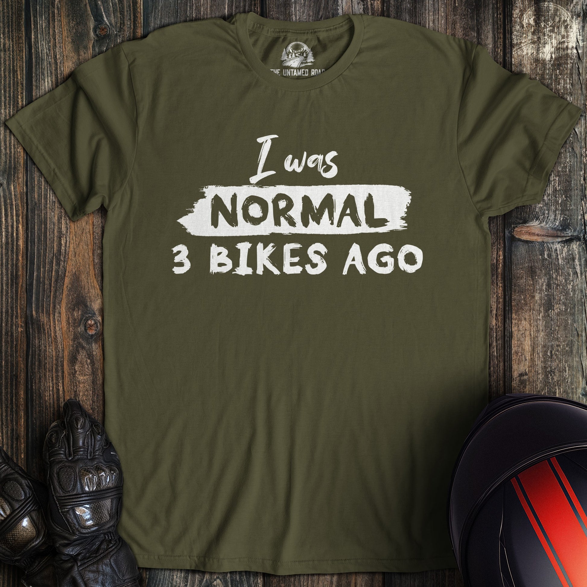 A military green colored t-shirt with the words "normal three bikes ago" printed on the front.