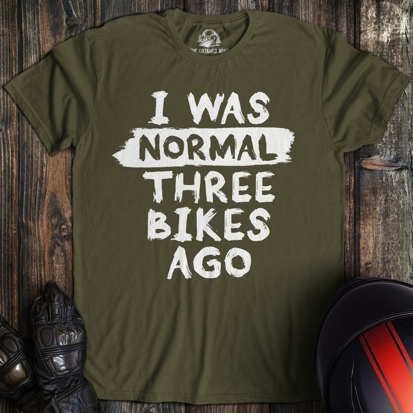 A military green colored t-shirt with the words "normal three bikes ago" printed on the front.