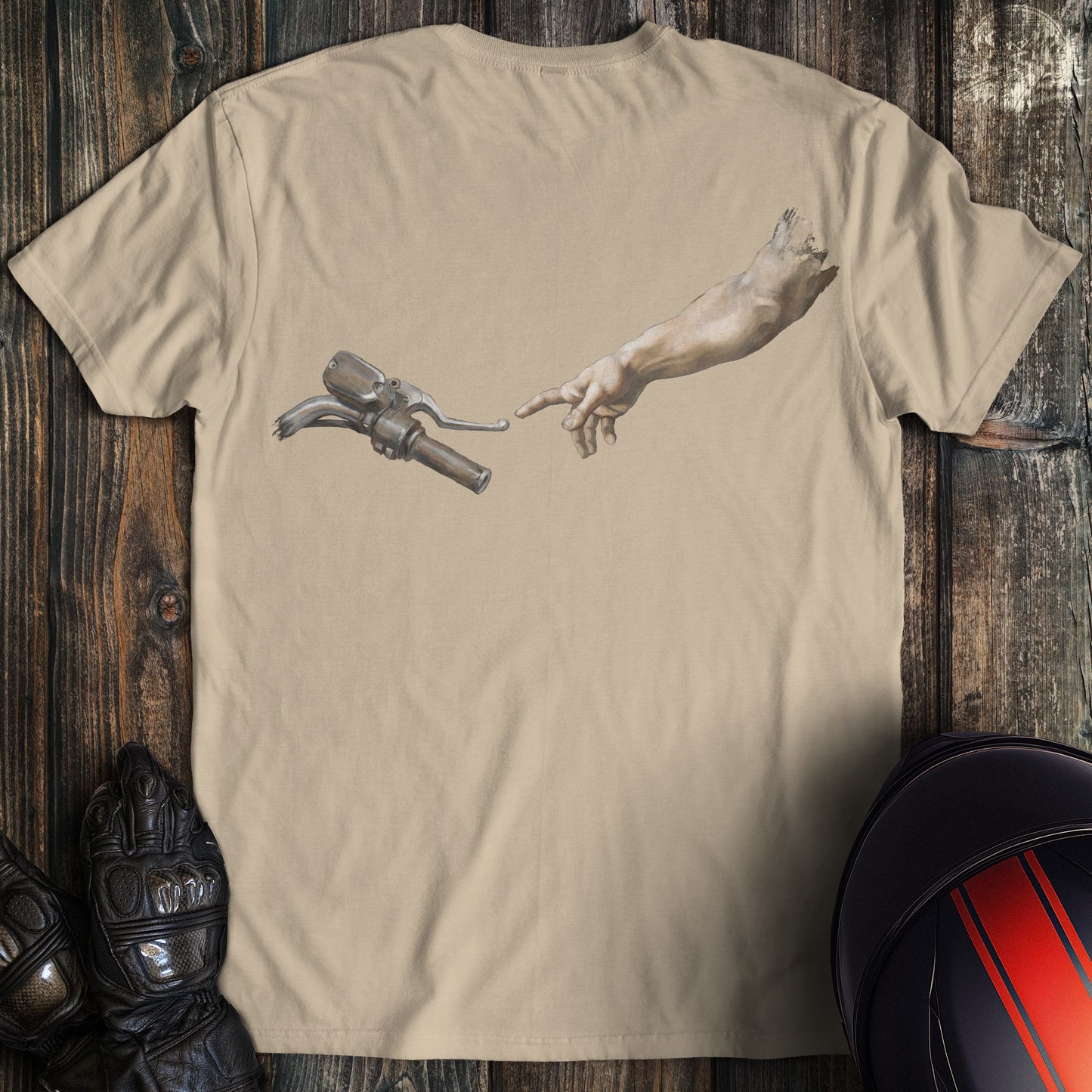 A sand colored t-shirt featuring a funny graphic of a hand reaching for a motorcycle handlebar in a creation style.