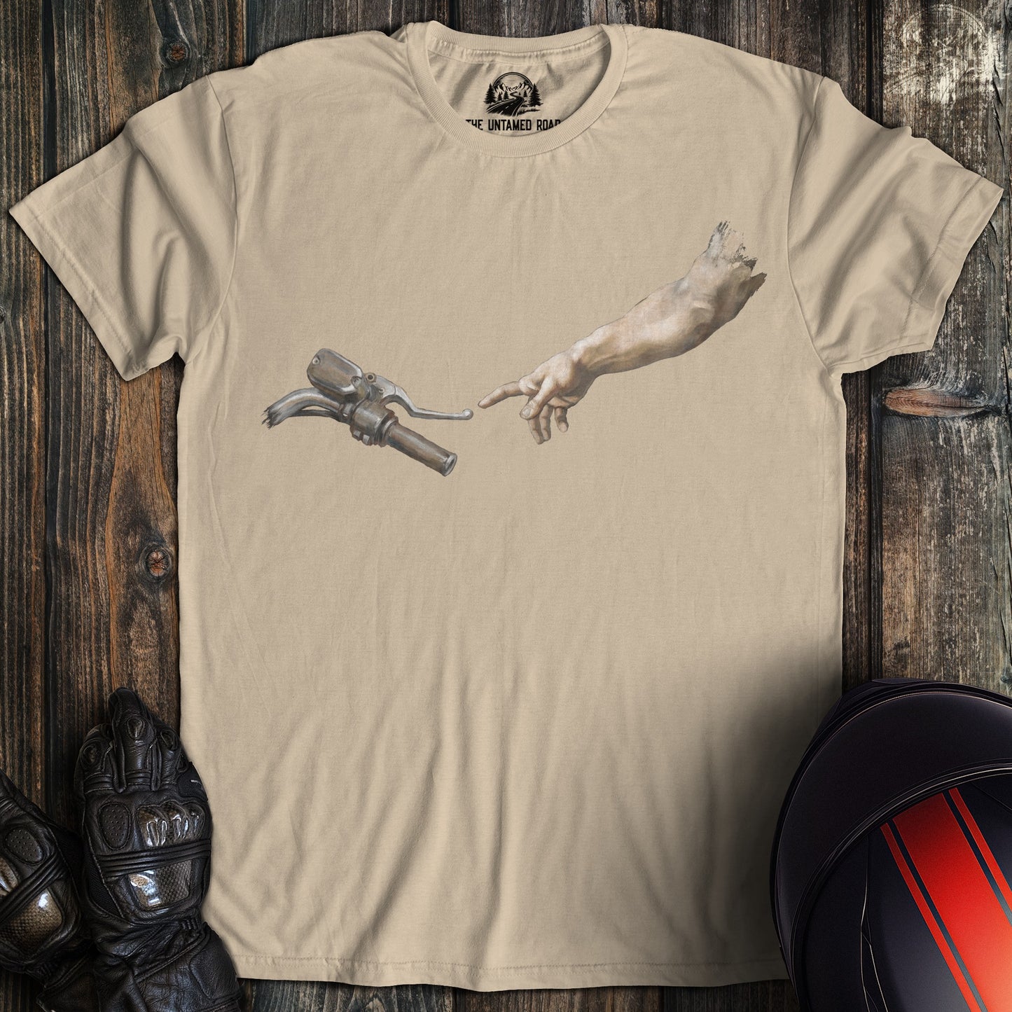 A sand colored t-shirt featuring a graphic of a hand reaching toward a motorcycle handlebar in a parody of the Creation.