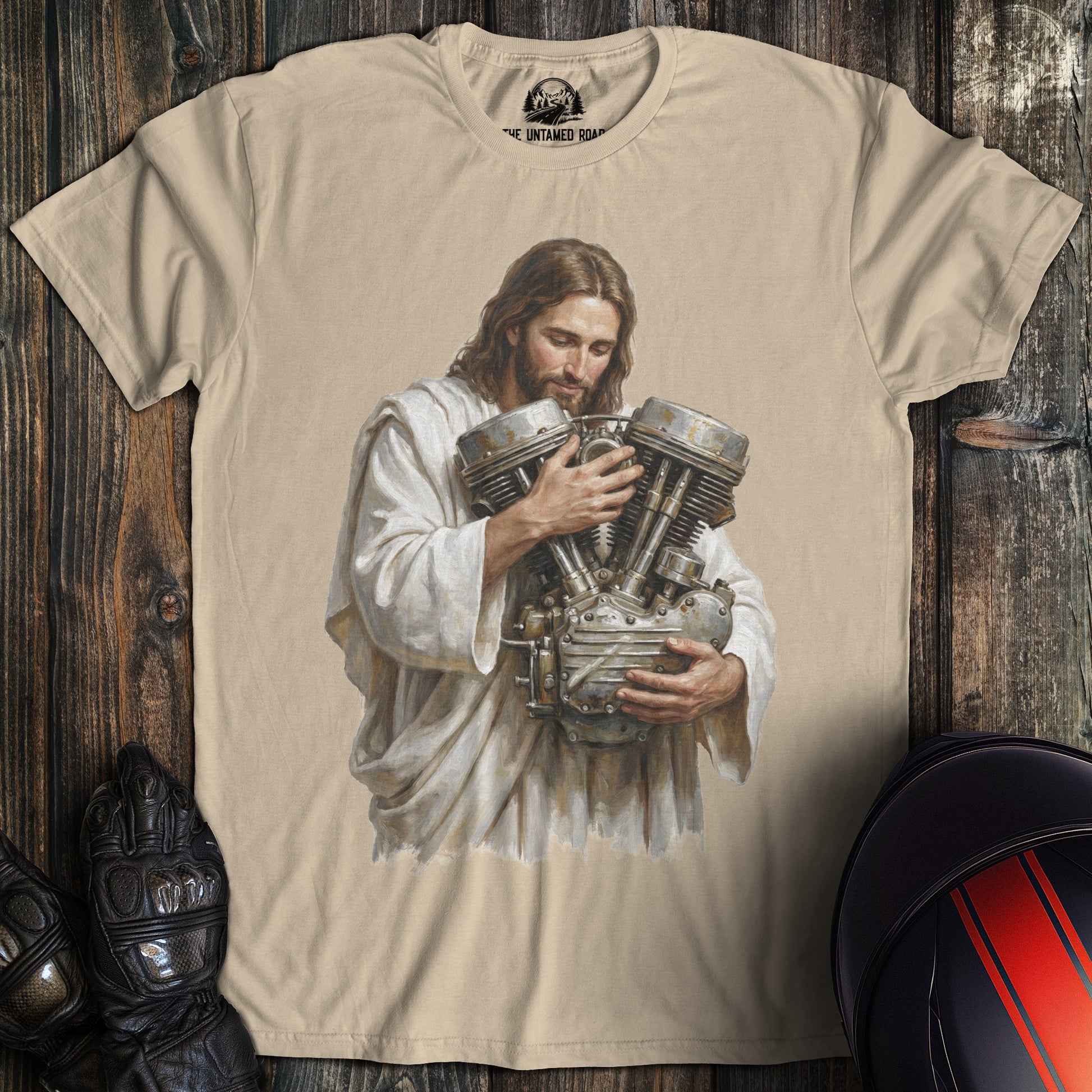 A sand colored t-shirt featuring a funny graphic of Jesus cradling a large motorcycle engine.